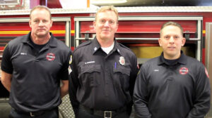 3 firemen stand smiling in front of fire engine